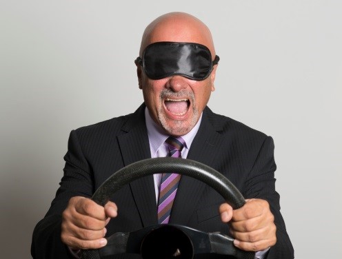A blindfolded man in a suit grips a steering wheel, shouting