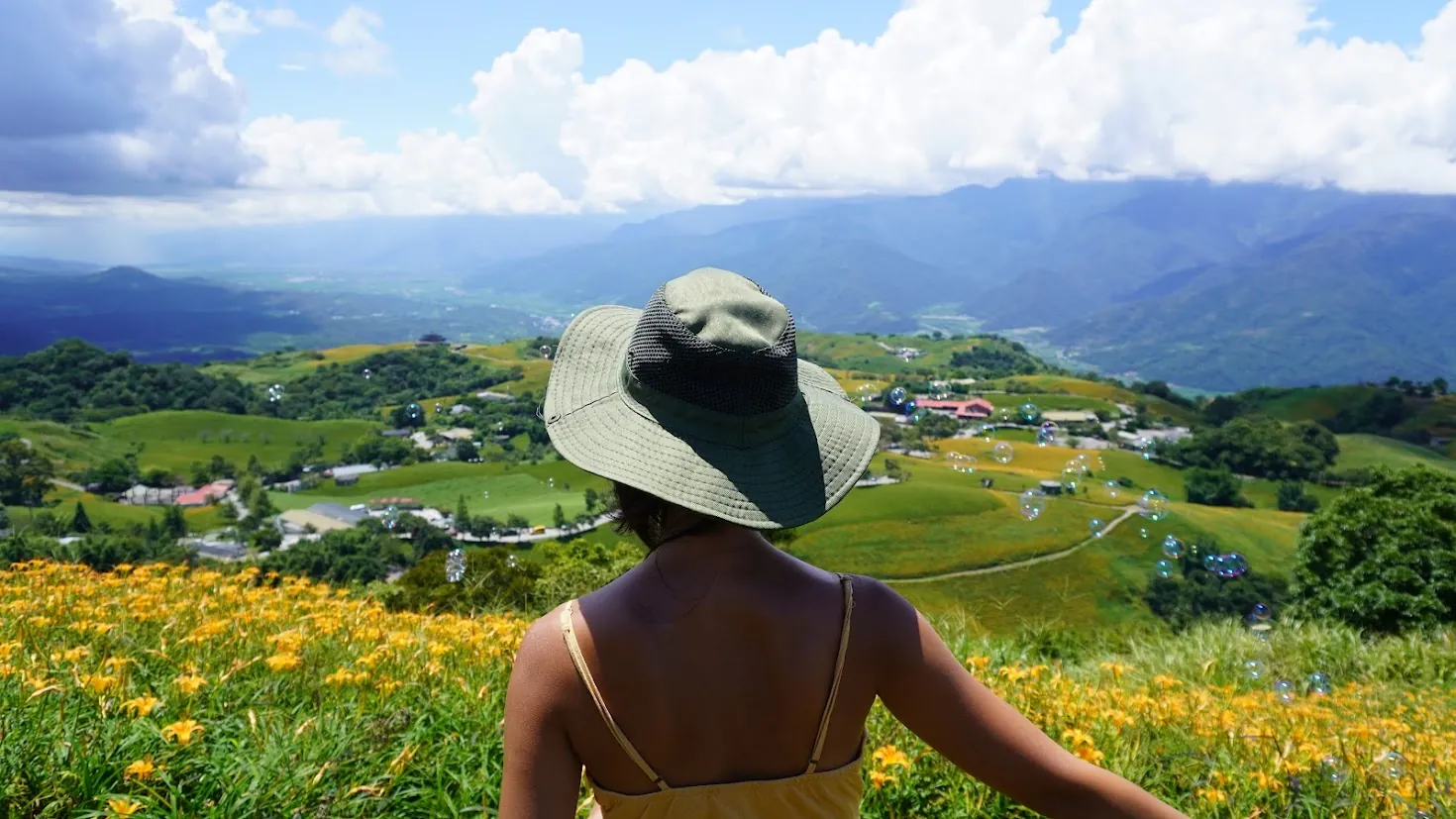 Fuli, Hualien: a woman in a sun hat looking out over golden needle flowers, green hills and mountains