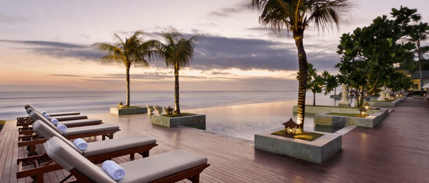A beach resort at sunset: loungers on a deck beside an infinity pool and palm trees facing the ocean