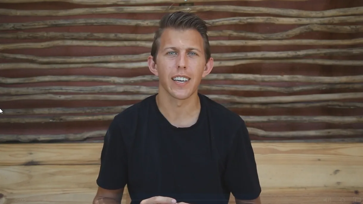 Video still: Paul talking to the camera in front of a wall of rough wooden poles