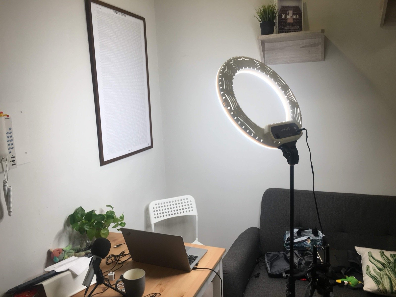 A small home studio: a ring light on a stand, a laptop and microphone on a wooden desk, a framed life calendar on the wall and a sofa
