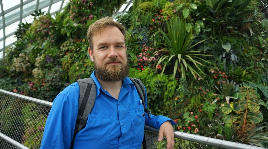 Artur Piszek in a blue shirt and backpack in front of a wall of tropical plants