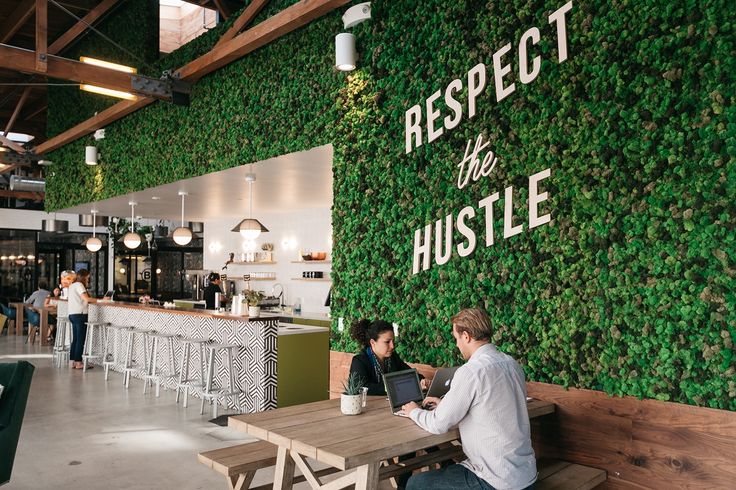 A WeWork lounge with a moss wall that reads Respect the Hustle and two people working on laptops at a wooden table