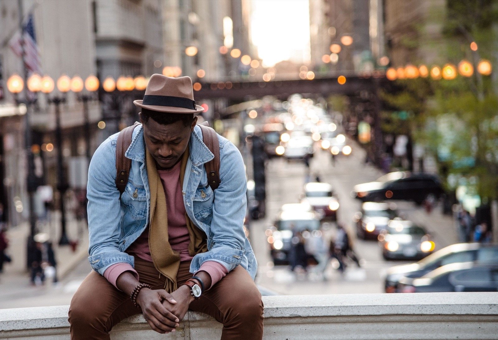 A man in a hat and denim jacket sits on a ledge above a busy city street, head bowed